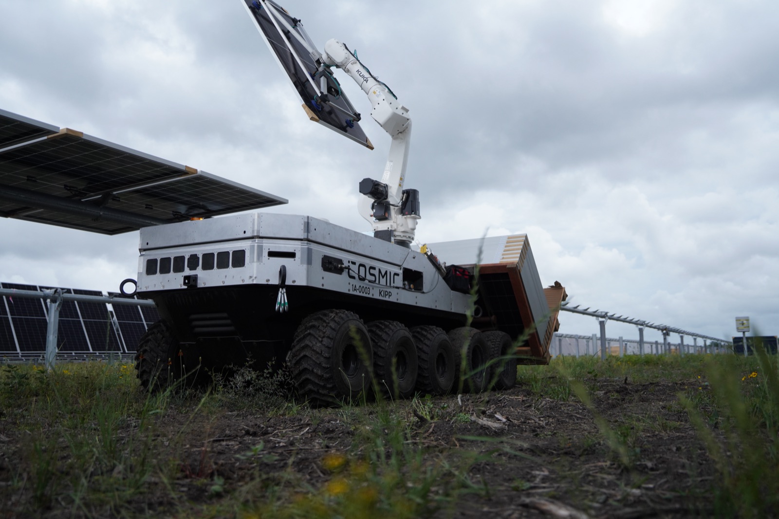 Cosmic-1 lifting a solar module overhead with its robotic arm at a tracker row under an overcast sky